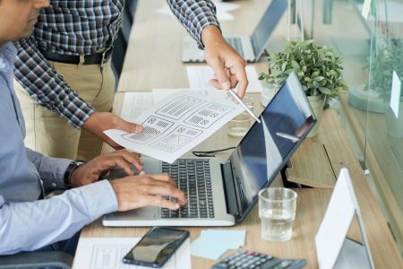 UX designers comparing sketch of interface with finished website design on laptop screen